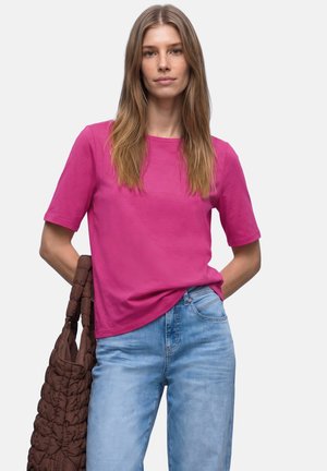 Mujer con cabello rubio largo que lleva una camiseta de manga corta color rosa brillante y unos vaqueros azul claro, sosteniendo un gran bolso tote marrón acolchado.