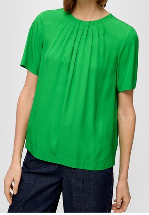 Woman wearing a bright green short-sleeve blouse with pleats at the neckline and dark blue jeans against a plain background.