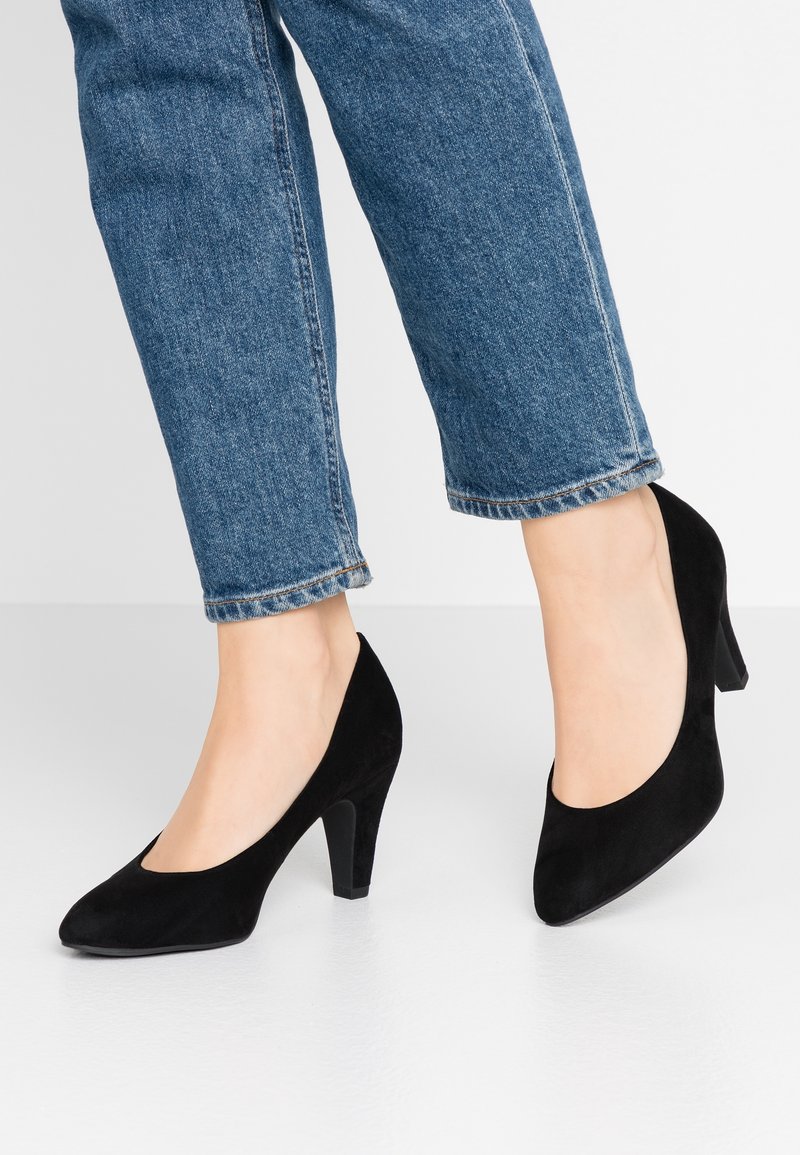 Chaussures à talons hauts en daim noir avec un bout pointu, présentant une texture lisse et un talon solide. Portées avec un pantalon en denim bleu court.