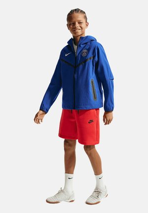 Jongen met gevlochten haar die een blauwe Paris Saint-Germain jas draagt, rode Nike shorts, witte Nike sokken en witte sportschoenen, staand en glimlachend.