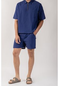 Námořnické pruhované tričko s krátkým rukávem a zipovým límečkem, doplněné o odpovídající šortky. Materiál má strukturovaný povrch. Šedé sandály dokreslují celý outfit.