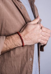 Brățară cu mărgele rotunde din lemn roșiatic pe un încheietură, asortată cu o cămașă din catifea de culoare maro și un tricou gri. Prim-planul arată textura și detaliile.