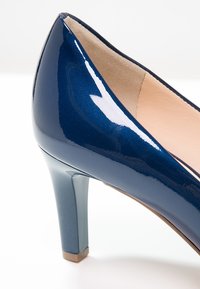 Marinblå lackläderpump med rundad tå och medelhög blockklack. Slät textur med glansig finish. Beige inredning.