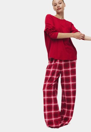 Joven con una camisa roja de manga larga y pantalones anchos de cuadros rojos y blancos, ajustándose la manga sobre un fondo blanco.