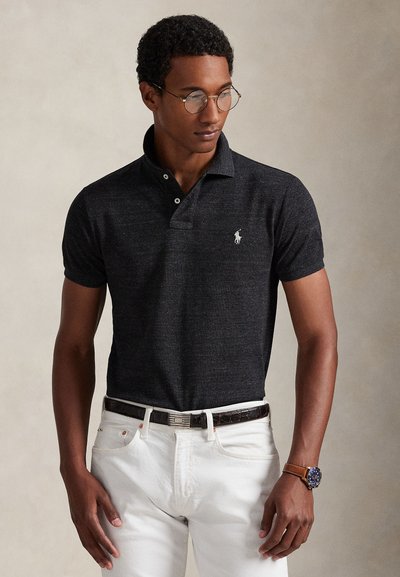 Young man wearing round glasses, dark short-sleeve polo shirt, white pants with black belt, and brown wristwatch, looking down to the side.