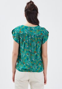 Grüne Blumenbluse mit kurzen Ärmeln und einem gerafften Rückendesign. Der Stoff zeigt bunte Pflanzenmuster auf einem strukturierten Hintergrund.