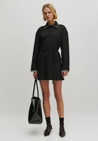 Schwarzes Hemdkleid mit langen Ärmeln, Knopfleiste, tailliertem Gürtel und kontrastierendem weißen Kragen. Das Model hält eine schwarze, glänzende Tragetasche.