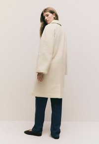Cream, textured wool-blend coat with a wide collar, long sleeves, and side pockets, paired with dark blue wide-leg jeans and flat shoes.