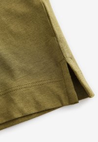 Gros plan de l'ourlet en tissu vert olive avec une double couture et une petite fente latérale sur un vêtement léger.