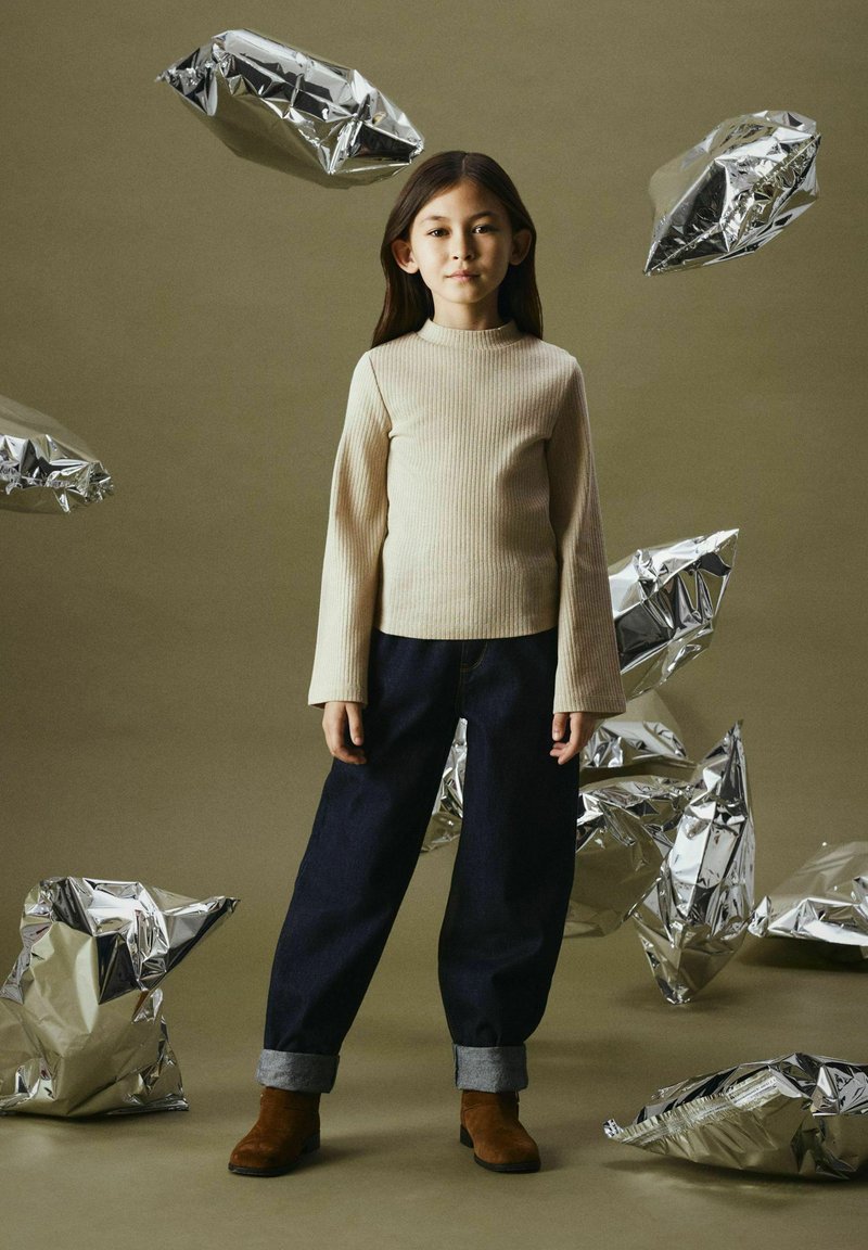 Beige ribbad långärmad topp, mörka denimvidbyxor med upprullade fållar, bruna ankelboots. Silverfärgade skrynkliga folieaccenter i bakgrunden.