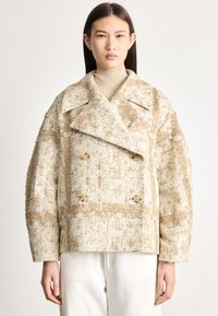 Sieviete, kas valkā bēša un baltas teksturētas divrindu vilnas mētelis pār bēša augstu pieskaņotu džemperi un baltas bikses, stāvot pret vienkāršu fonu.