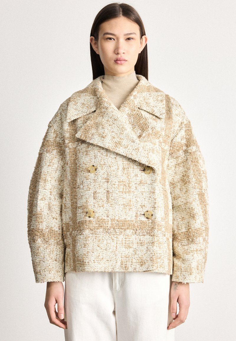 Sieviete, kas valkā bēša un baltas teksturētas divrindu vilnas mētelis pār bēša augstu pieskaņotu džemperi un baltas bikses, stāvot pret vienkāršu fonu.