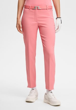 Eng geschnittene rosa Hose mit passendem Gürtel, kombiniert mit weißen Turnschuhen und einem weißen Golfhandschuh an der rechten Hand.