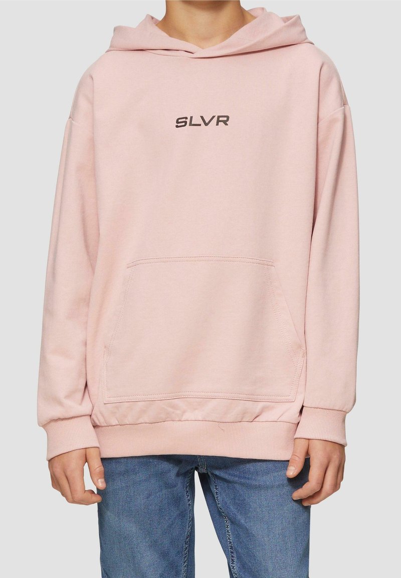 Personne portant un sweat à capuche rose pâle avec poche avant et texte noir "SLVR", associée à un jean en denim bleu sur un fond uni.