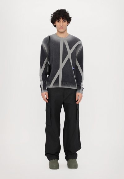 Pullover in verlaufendem Grau mit geometrischen hellgrauen Mustern, kombiniert mit schwarzen Cargo-Hosen und grünen Slippern. Schlichte schwarze Tasche.