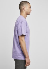 Tricou din bumbac violet cu mânecă scurtă, guler tip crew și croială lejeră, prezentat din lateral, asortat cu pantaloni gri închis.