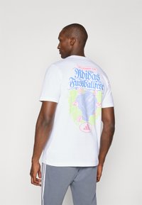 Hvid bomulds t-shirt med et farverigt grafisk motiv på ryggen, som har et fodbolddesign, tekst i blå og pink samt Adidas-logoet nederst.