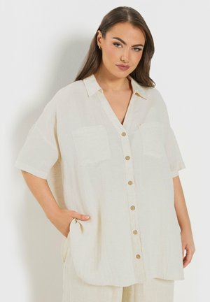 Femme portant une chemise ample, boutonnée écrue avec poches poitrine, debout avec une main dans la poche devant un fond clair uni.