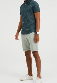 Marinblå kortärmad skjorta med knappar och geometriskt mönster, kombinerad med ljusgröna shorts. Accessoarer inkluderar en klocka och vita sneakers.