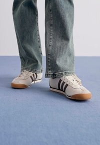 Piedi che indossano sneaker bianche con strisce nere e suole in gomma, poggiati su un pavimento blu, con jeans blu larghi che coprono le caviglie.
