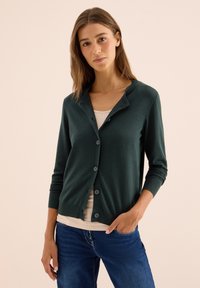 Junge Frau mit langen braunen Haaren, die eine dunkelgrüne, mit Knöpfen versehene Jacke, ein beiges Oberteil und blaue Jeans trägt, posiert mit einer Hand in der Tasche.