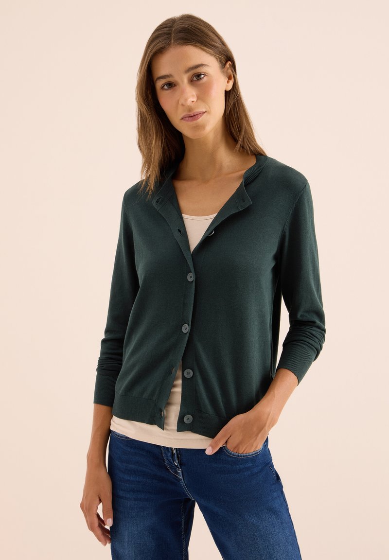 Junge Frau mit langen braunen Haaren, die eine dunkelgrüne, mit Knöpfen versehene Jacke, ein beiges Oberteil und blaue Jeans trägt, posiert mit einer Hand in der Tasche.