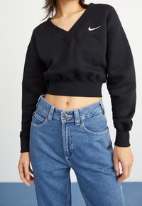 Svart croppad sweatshirt med V-ringning och ribbad nederkant, med en vit Nike-logga. Matchad med högmidjade blå denimjeans.