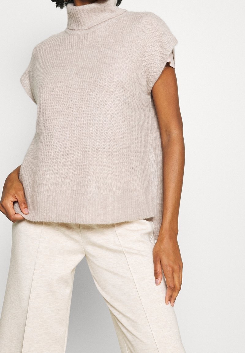 Beige gebreide mouwloze coltrui met een ontspannen pasvorm, voorzien van een geribbelde textuur en een rechte zoom. Gecombineerd met lichte broek.