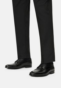 Fekete bőr alkalmi cipők sima felülettel, hegyes orral és klasszikus fűzős kivitelben, melyekhez testre szabott fekete nadrág társul.