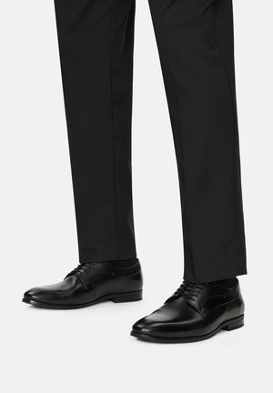 Juodos odinės suknelės bateliai su lygia apdaila, turintys smailų pirštą ir klasiką raištelių dizainą, derinami su pritaikytomis juodomis kelnėmis.