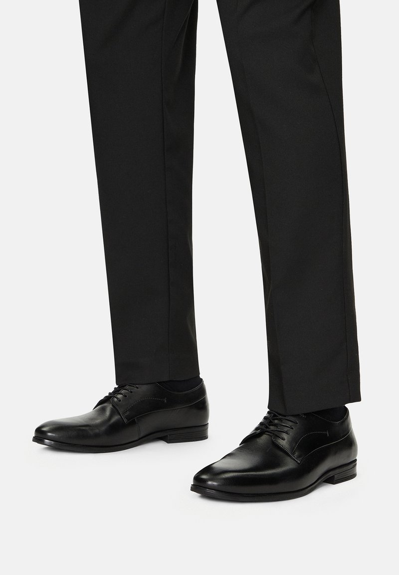 Fekete bőr alkalmi cipők sima felülettel, hegyes orral és klasszikus fűzős kivitelben, melyekhez testre szabott fekete nadrág társul.