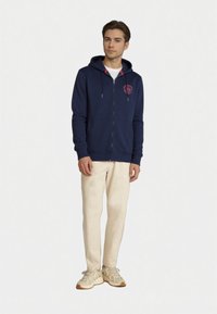 Felpa con zip colore blu navy, con un logo rosa, dotata di cappuccio con cordino, tasche frontali e polsini a coste, indossata con pantaloni beige chiaro e sneakers color cammello.