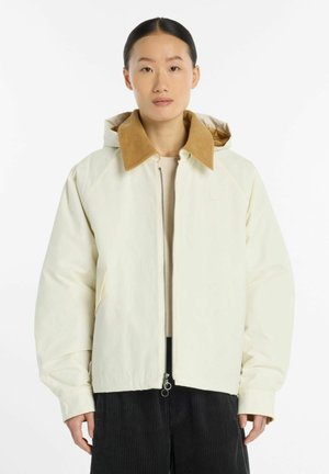Person står med fronten mod kameraet iført en offwhite jakke med en lysebrun fløjlskrave og sorte bukser mod en ensfarvet hvid baggrund.