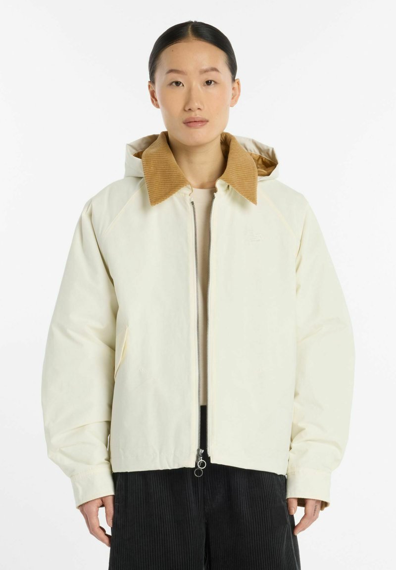 Personne debout de face, portant une veste écrue au col en velours côtelé beige et un pantalon noir sur fond blanc uni.