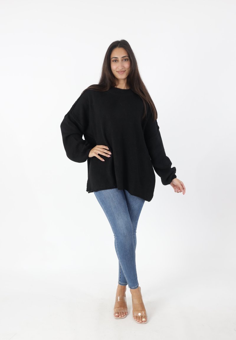 Maglione nero oversize a maglia con maniche lunghe, abbinato a jeans skinny blu chiaro e sandali con tacco trasparente, su uno sfondo neutro.