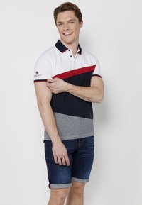 Tricou polo alb cu dungi bleumarin și roșii, cu un fund gri. Mâneci scurte și un guler cu două butoane. Asortat cu șorturi din denim întunecat, răsfrânte.