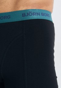 Black boxer briefs with a teal waistband featuring the brand name "BJÖRN BORG" in dark blue. Made from smooth, stretchy fabric.