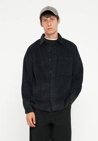 Black checkered button-up shirt with a collar, made of a textured fabric, worn over a black shirt. Paired with a light-colored cap.