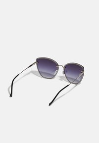 Gafas de sol estilo cat-eye con lentes gradientes en color púrpura, marco de metal en tono plateado y detalles decorativos de cadena a lo largo de los bordes de los lentes. Patillas negras.