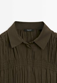 Chemise col olive foncé avec plis verticaux et détail de couture horizontale sur la poitrine, étiquette indiquant "Massimo Dutti, Made in China."