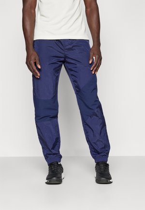 Pantalons de sport légers bleu marine avec une texture brillante, présentant une coupe ajustée et des poignets élastiques, associés à des baskets noires.