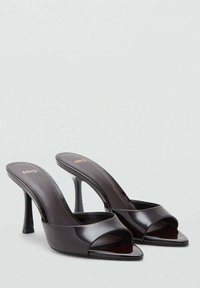 Los mules de tacón alto en color negro brillante tienen la puntera abierta, amplias correas frontales y un diseño de tacón curvado y elegante. Marca sutil en las plantillas.