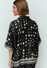 Schwarze Satinbluse im Oversized-Schnitt, mit weißen und goldenen Punkten. Sie hat einen klassischen Kragen und ein gestreiftes Saumdetail.