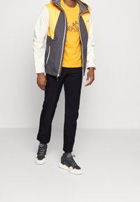 Kurtka z blokami kolorów w odcieniach pomarańczowym, szarym i białym; żółta koszula z czarnym logo; czarne spodnie; szaro-czarne sneakersy z białymi podeszwami.