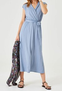 Ljusblå omslagsklänning med korta ärmar, knytbälte vid midjan och knälång nederkant. Modellen håller ett sjal med blommönster.