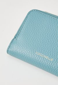 Carteira de pele verde-água com superfície texturizada, logótipo dourado e fecho zip, com bordas arredondadas e design minimalista.