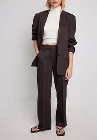 Femme portant un blazer et un pantalon marron foncé avec un pull à col roulé blanc raccourci et des sandales noires à bout carré, se tenant devant un fond uni.