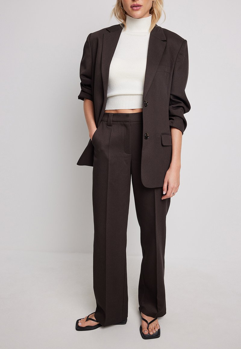 Femme portant un blazer et un pantalon marron foncé avec un pull à col roulé blanc raccourci et des sandales noires à bout carré, se tenant devant un fond uni.