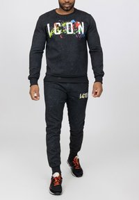 Ensemble de sport noir en tissu texturé ; présente un motif de taches colorées sur le logo. Poignets et taille côtelés ; associé à des baskets noires.