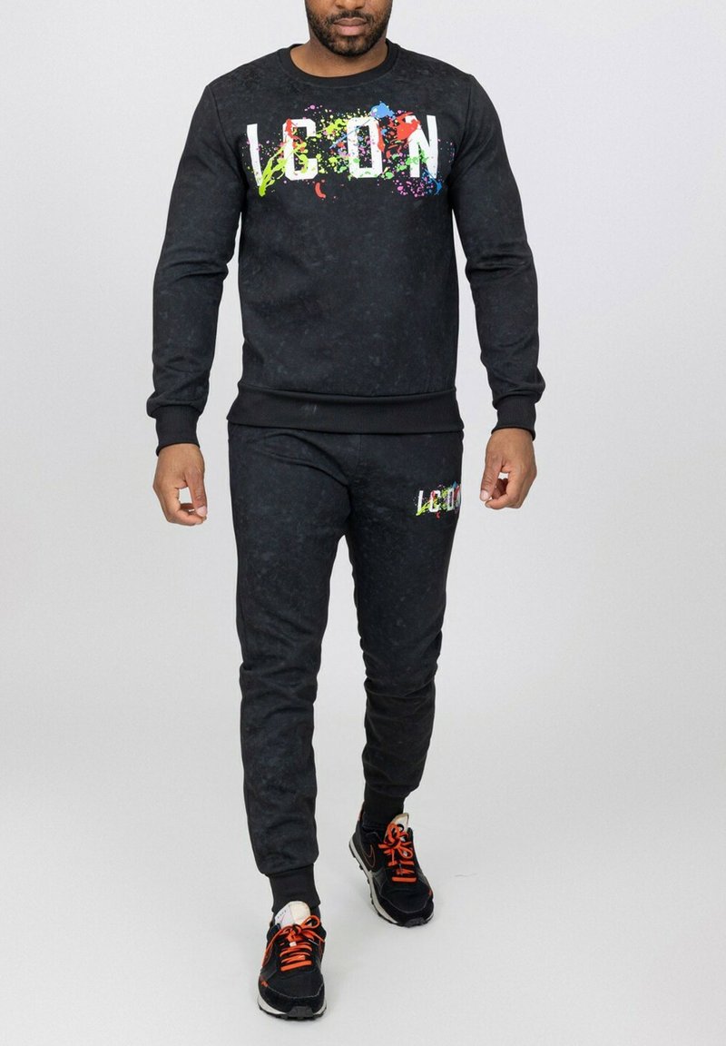 Ensemble de sport noir en tissu texturé ; présente un motif de taches colorées sur le logo. Poignets et taille côtelés ; associé à des baskets noires.